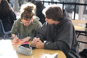  Working together, senior Bannon McCleve helps junior Emmy Hedges with writing a letter to senior citizens in nursing homes. These letters provided an opportunity to brighten the lives of those who may feel isolated or lonely. “With the goal of spreading love and positivity to those who may not often have the chance to visit family,” McCleve said. “Our aim was to bring some kindness and joy to their lives, letting them know they are remembered and appreciated.”
Photo Credit: Achol Akot 