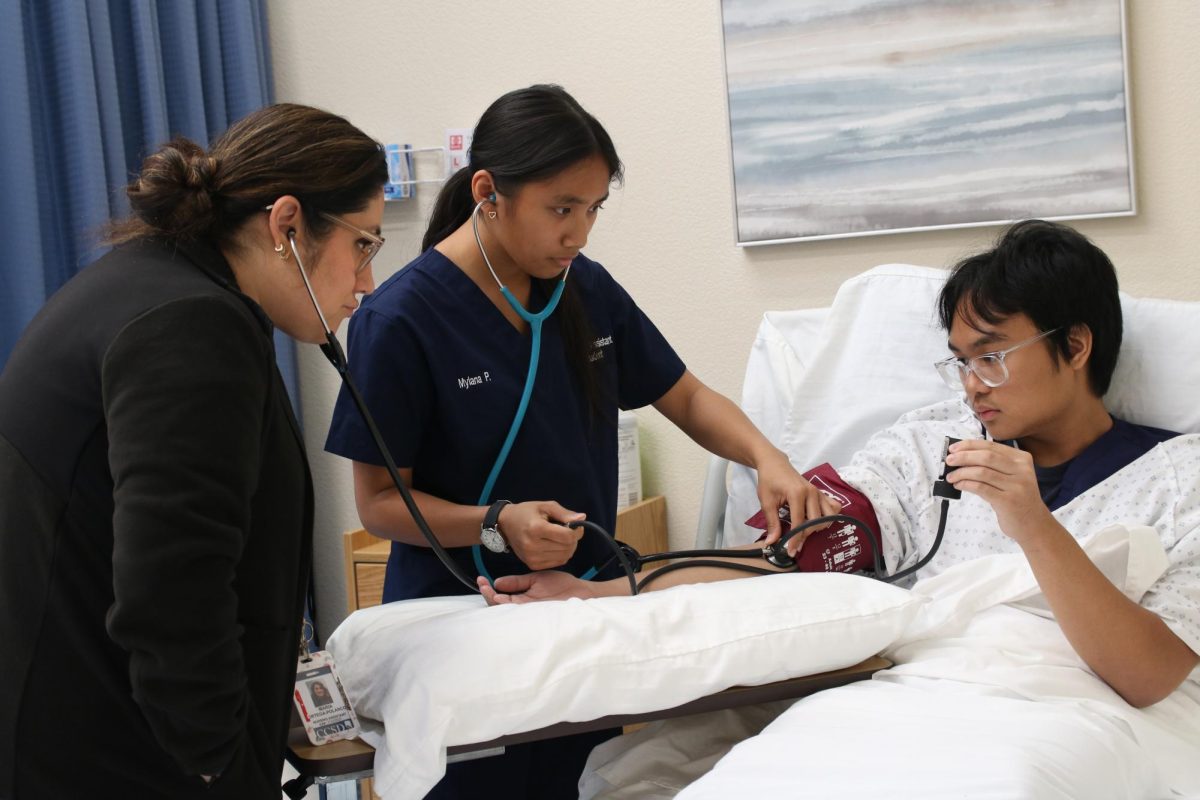 Focused on her patient, Maylana Param performs manual blood pressure on Jett Halili while Nursing teacher Maria Ortega Polanco verifies the results.